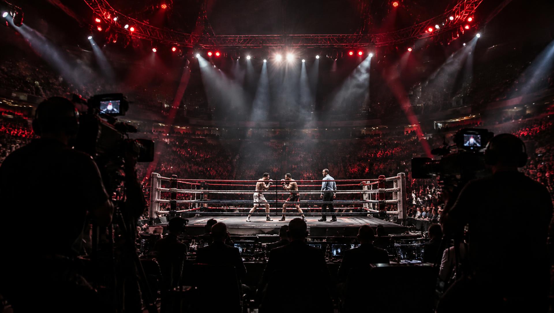 Ringside broadcast cameras filming a boxing bout in a packed arena